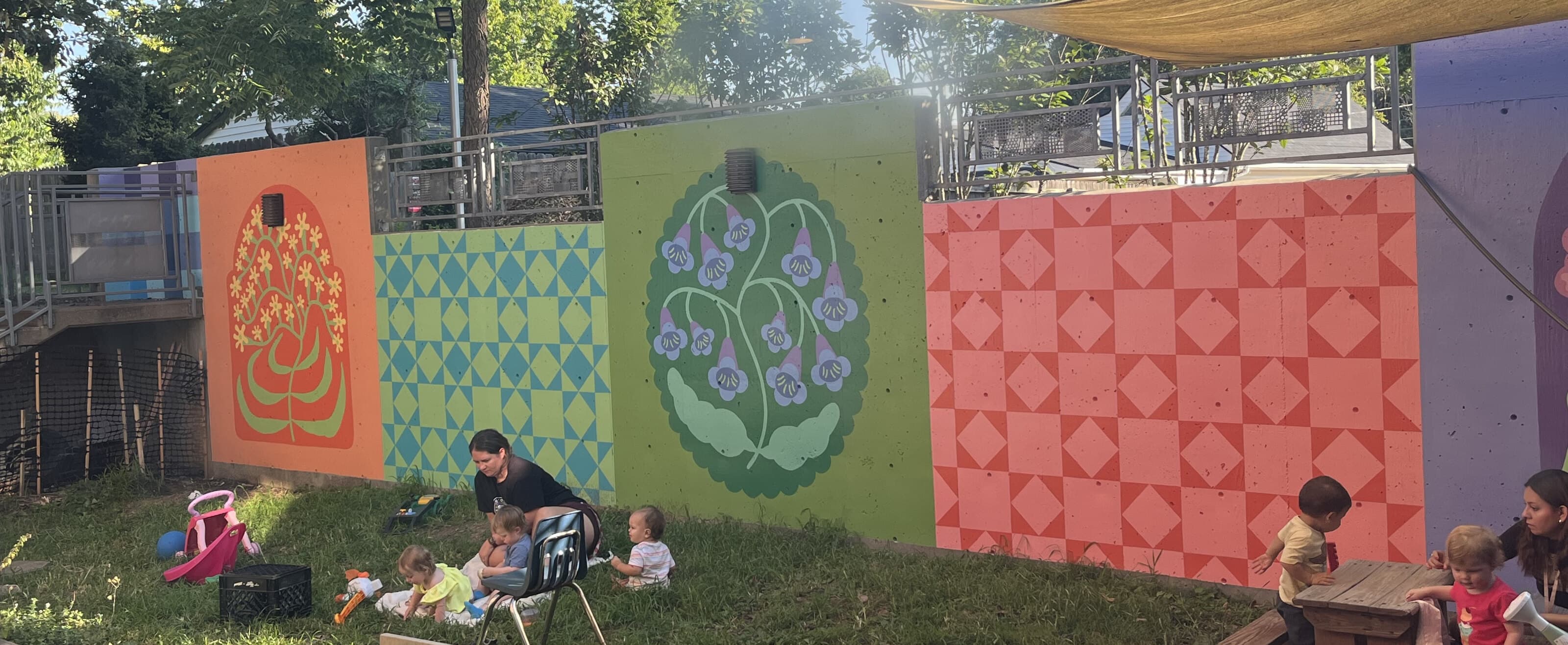A teacher and children on the grass beside the hand-painted mural wall at Lexington Friends Preschool